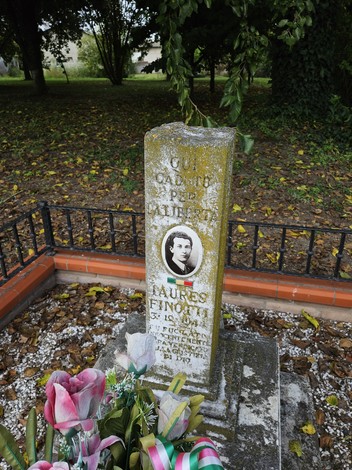 Cippo commemorativo di Finotti, con fiori e decorazioni, immerso nel verde e circondato da una recinzione.