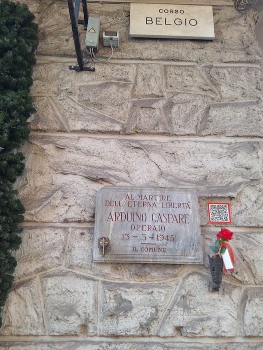 Lapide in marmo su parete in pietra, con iscrizione e fiore in un vaso tricolore sotto.
