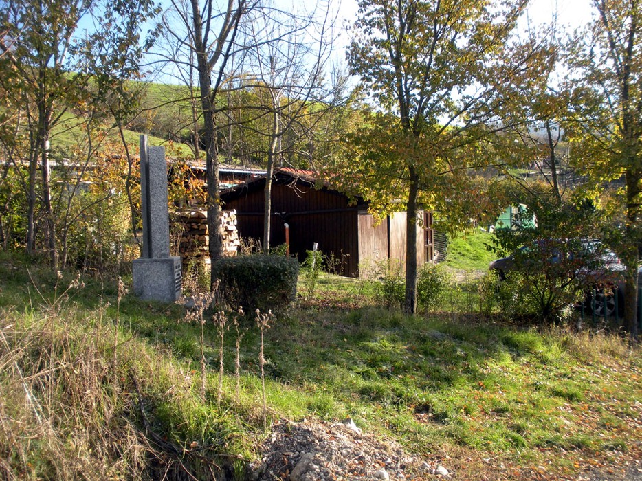 Cippo in una zona verde con alberi, vicino a un edificio rustico e a materiali da costruzione.