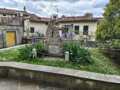 Monumento commemorativo circondato da piante e fiori, con edifici storici sullo sfondo.