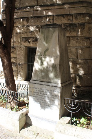 Cippo commemorativo a Pacifici, inciso con testi e situato in un'area urbanizzata con vegetazione circostante.