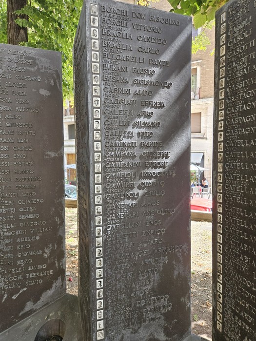 Monumento in lastre di pietra scura con iscrizioni, disposto all’aperto tra alberi e abitazioni.
