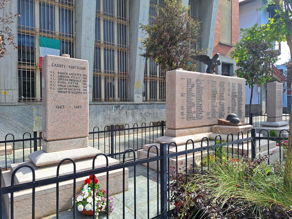 Monumento ai caduti, con iscrizioni commemorative e decorazioni floreali, circondato da una recinzione.