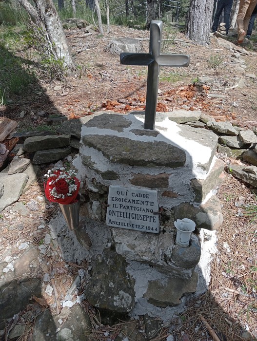 Cippo commemorativo di Ontelli Giuseppe, con una croce, fiori e candele, immerso nella natura.