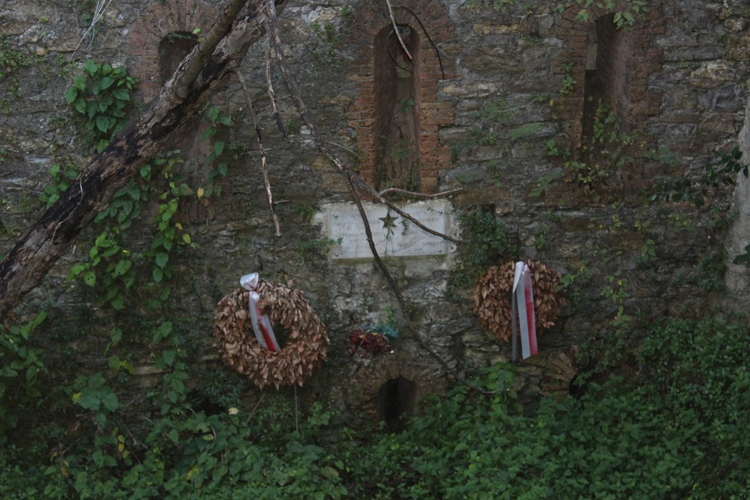 Lapide commemorativa con ghirlande e decorazioni, immersa nella vegetazione di un muro storico.