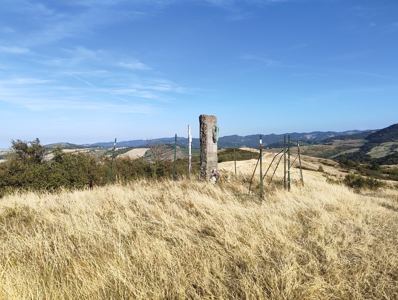 Stele in pietra con iscrizione, isolata tra l’erba alta su un crinale collinare.