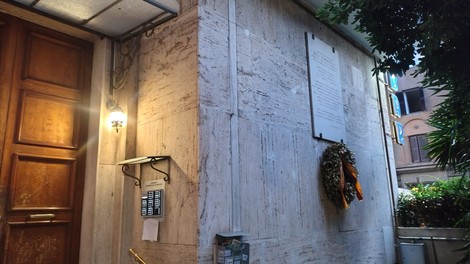 Lapide commemorativa su una parete, con una corona di fiori e un ingresso in legno sullo sfondo.