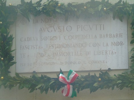 Lapide commemorativa in onore di Augusto Picutti, decorata con alloro e nastro tricolore.