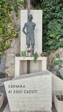 Monumento ai caduti, statua in bronzo affiancata da piante e scritte commemorative in un contesto naturale.