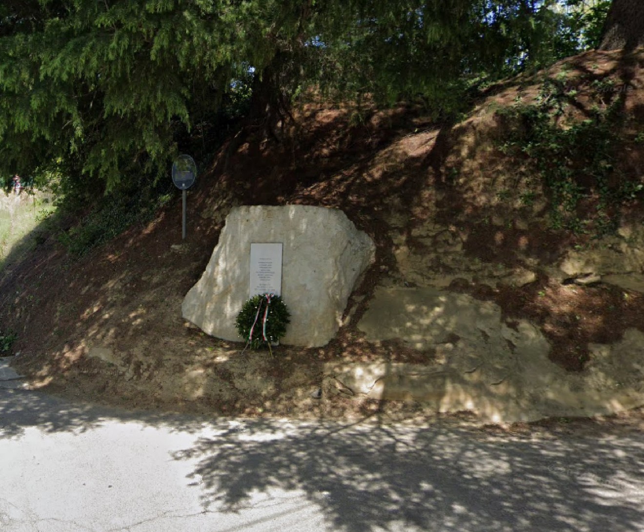 Lapide commemorativa incastonata in una roccia, circondata da vegetazione e decorata con una corona di fiori.