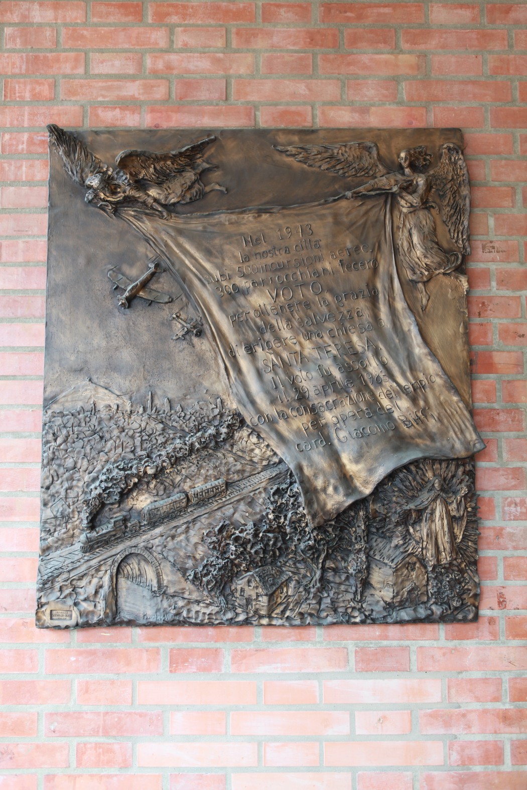 Rappresentazione bronzea di un ex-voto dedicato alle vittime dei bombardamenti, con figure angeliche e scene di lotta.