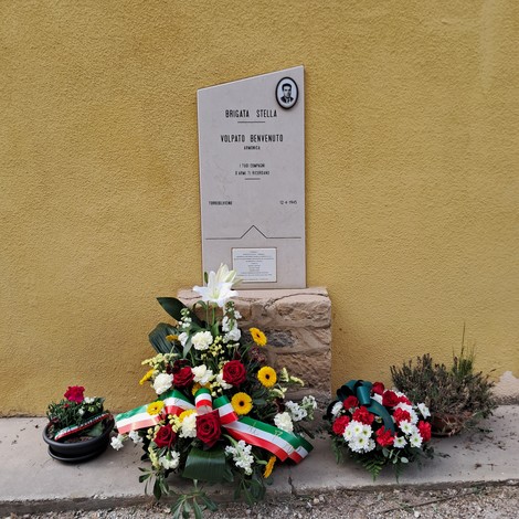 Lapide commemorativa dedicata a Volpato, con fiori e tributi nei pressi di una parete gialla.
