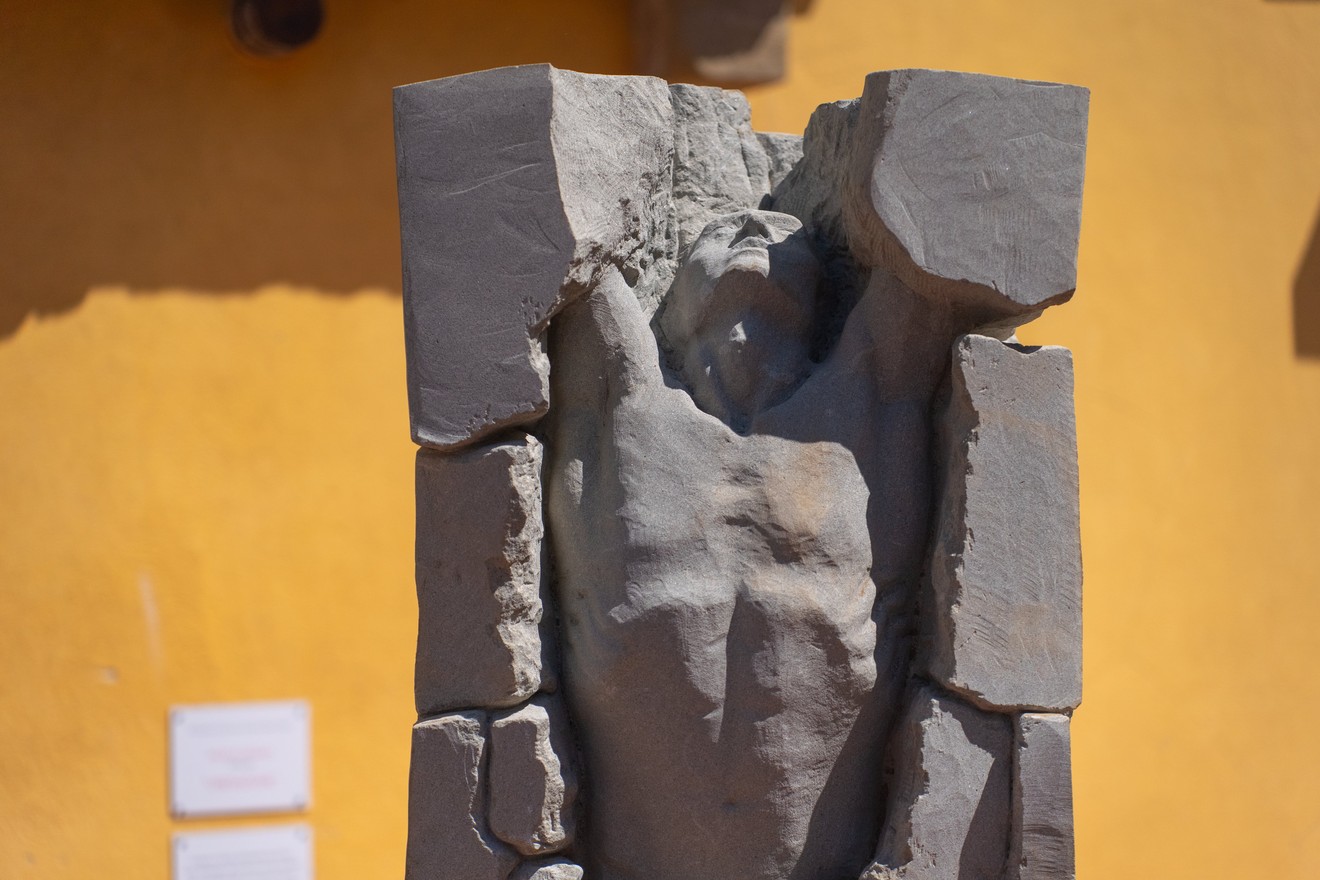 Statua in pietra grigia con figura scavata al centro, su sfondo di parete gialla.