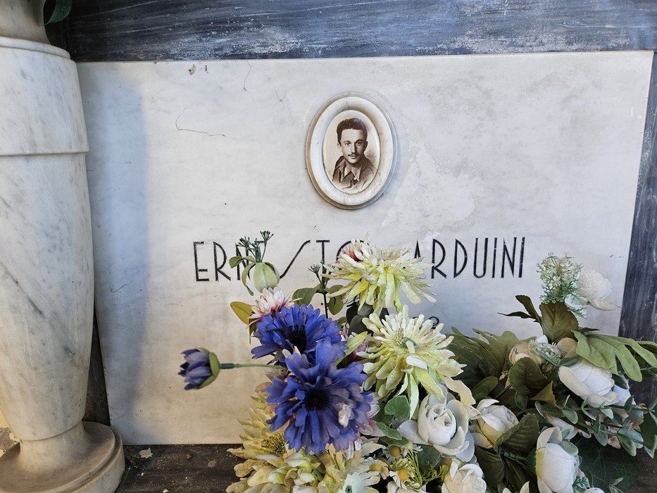 Lapide in marmo con ritratto ovale e iscrizione, ornata da fiori deposti davanti.