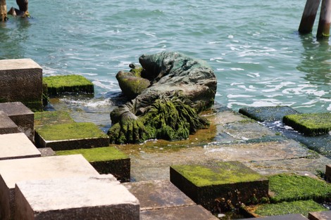 Monumento alla partigiana immerso nella natura, con vegetazione che cresce attorno alla statua e acqua sullo sfondo.