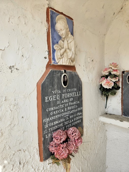 Lapide in pietra con immagine sacra, ritratto e iscrizione, su parete esterna con fiori decorativi.