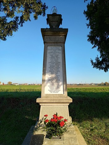 Monumento ai caduti dell'Amola, con iscrizioni e fiori rossi, immerso in un paesaggio sereno e verdeggiante.