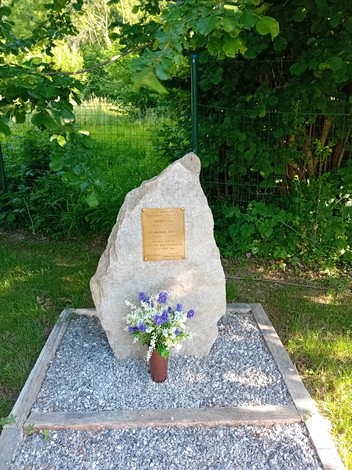 Cippo commemorativo con fiorellini, circondato da verde e pietre, in un'area tranquilla e naturale.