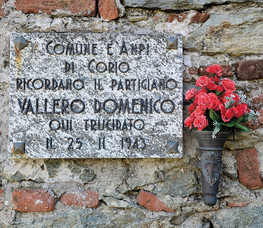 Lapide in marmo su muro di pietra, con iscrizione e vaso di fiori rossi accanto.
