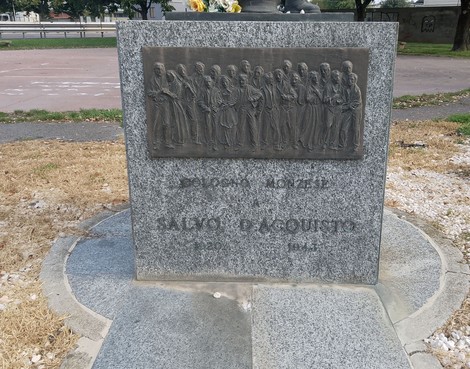 Monumento a D'Acquisto, commemorazione con rilievi bronzei su base in granito, situato in un’area verde.