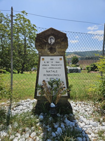 Cippo commemorativo dedicato a Natale Romagnoli, con fiori e paesaggio verde circostante.