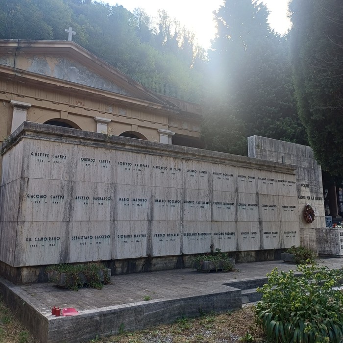 Sacrario in pietra con lapidi incise disposte su una parete, davanti a un edificio e alla vegetazione.