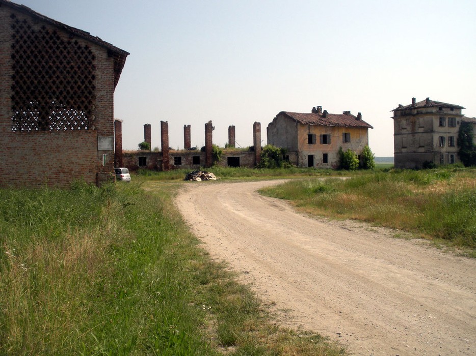 Area abbandonata con edifici in rovina e un terreno erboso che circonda una strada sterrata.