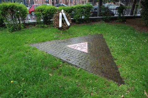 Monumento dedicato alle vittime omosessuali, con base triangolare e decorazioni floreali nel verde circostante.