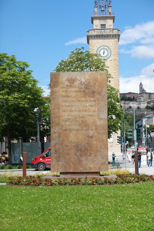 Monumento al partigiano, stele commemorativa con iscrizioni su sfondo verde e architettura cittadina.