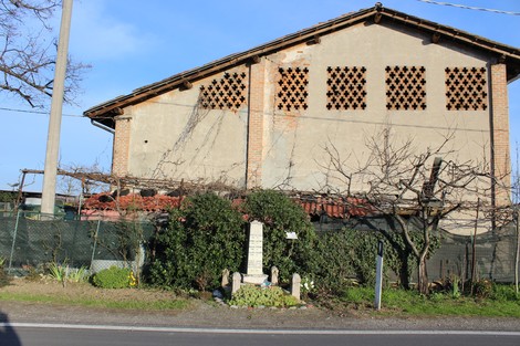 Monumento ai caduti dell'Insurrezione, circondato da vegetazione, con edificio storico sullo sfondo.
