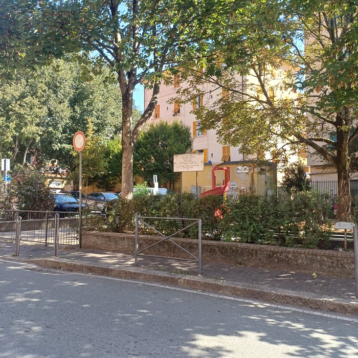 Via alberata con area verde recintata, edifici sullo sfondo; la lapide non è chiaramente visibile.
