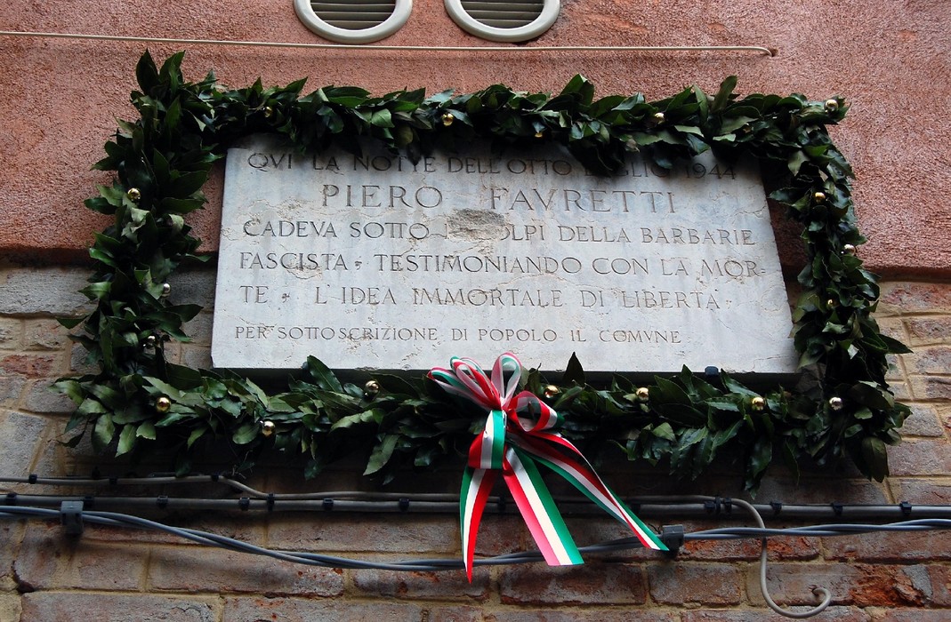Lapide commemorativa dedicata a Piero Favretti, circondata da una coroncina di foglie e decorazioni colorate.