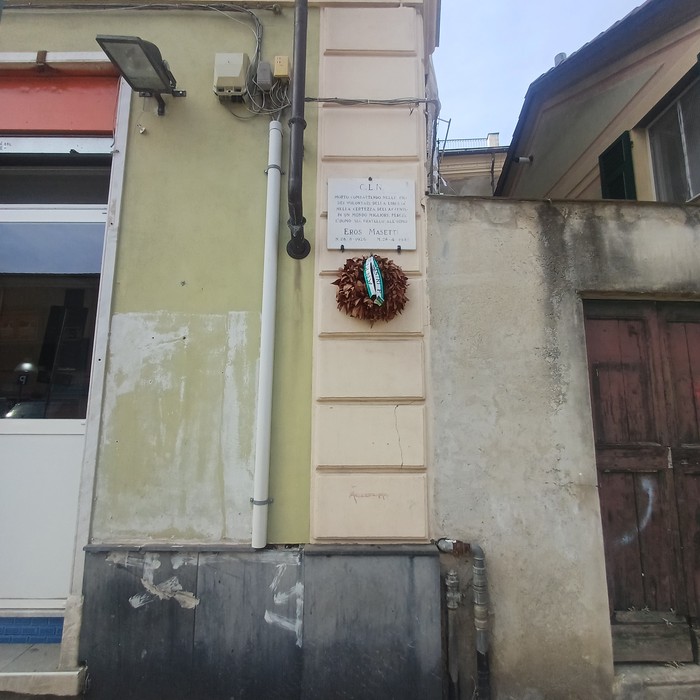 Lapide con iscrizione su parete esterna, sotto una composizione decorativa a forma di foglia.
