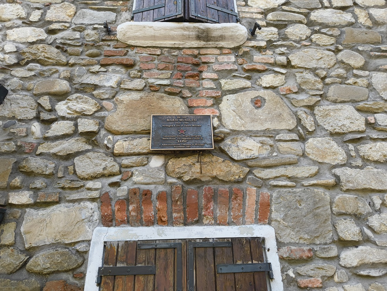 Targa su muro in pietra, dedicata a Musolesi, con finestre chiuse e dettagli in mattoni rossi.
