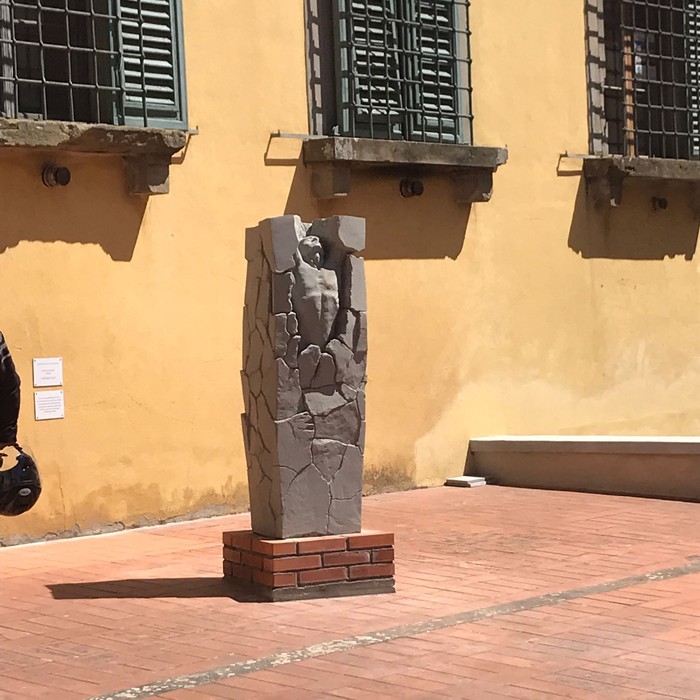 Statua in pietra grigia con figura umana scolpita, su base in mattoni, in un cortile davanti a una parete gialla.