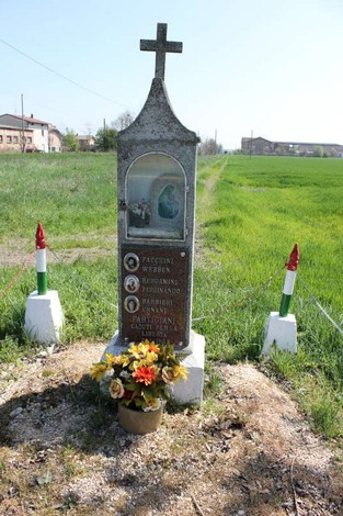 Tabernacolo monumentale con decorazioni floreali, situato in un paesaggio rurale, circondato da erba verde.