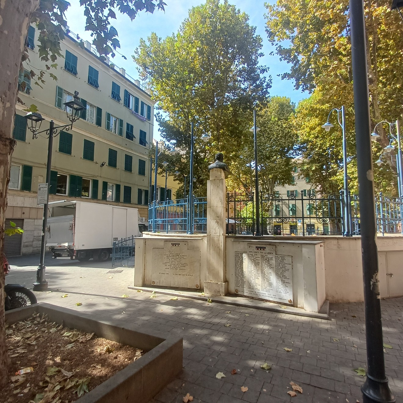 Monumento in pietra con lapidi incise, recintato, in una piazza alberata circondata da edifici.
