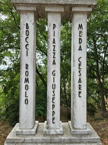 Cippo commemorativo con colonne, nomi incisi, circondato da vegetazione. Una testimonianza storica nel paesaggio.