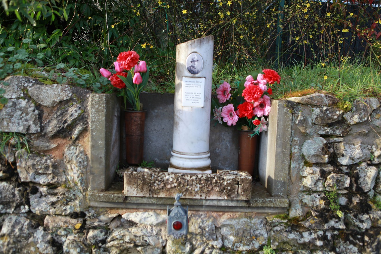 Cippo memoriale a Valgimigli con fiori colorati e decorazioni su un muro di pietra.