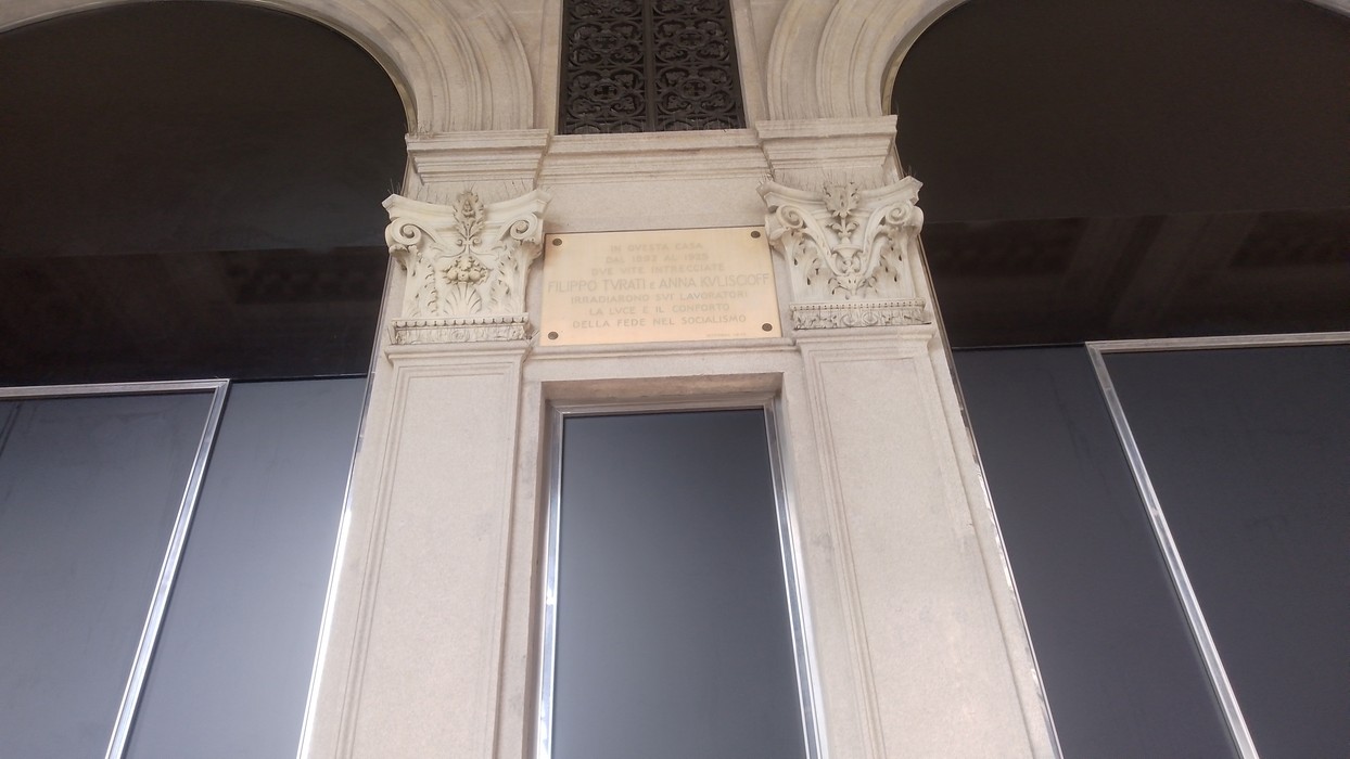 Targa con iscrizione tra due colonne scolpite, sopra un portone, su parete in marmo chiaro.