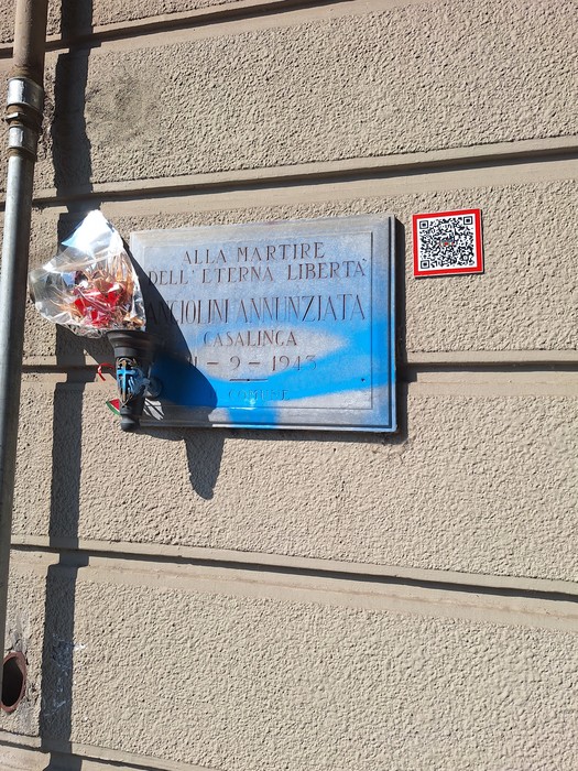 Lapide in marmo con iscrizione su parete esterna, con mazzo di fiori appeso sotto.