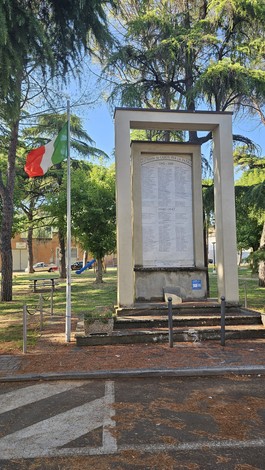 Monumento ai caduti circondato da alberi e una bandiera italiana, con una grande stele commemorativa.