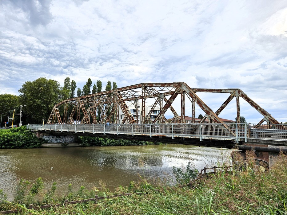 Ponte ferroviario in ferro arrugginito su un fiume, con sponde erbose in primo piano.