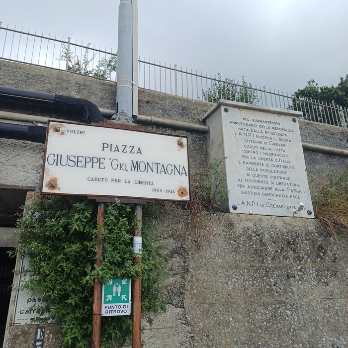 Targa con intitolazione della piazza e lapide con iscrizione affisse su un muro, sotto una ringhiera.