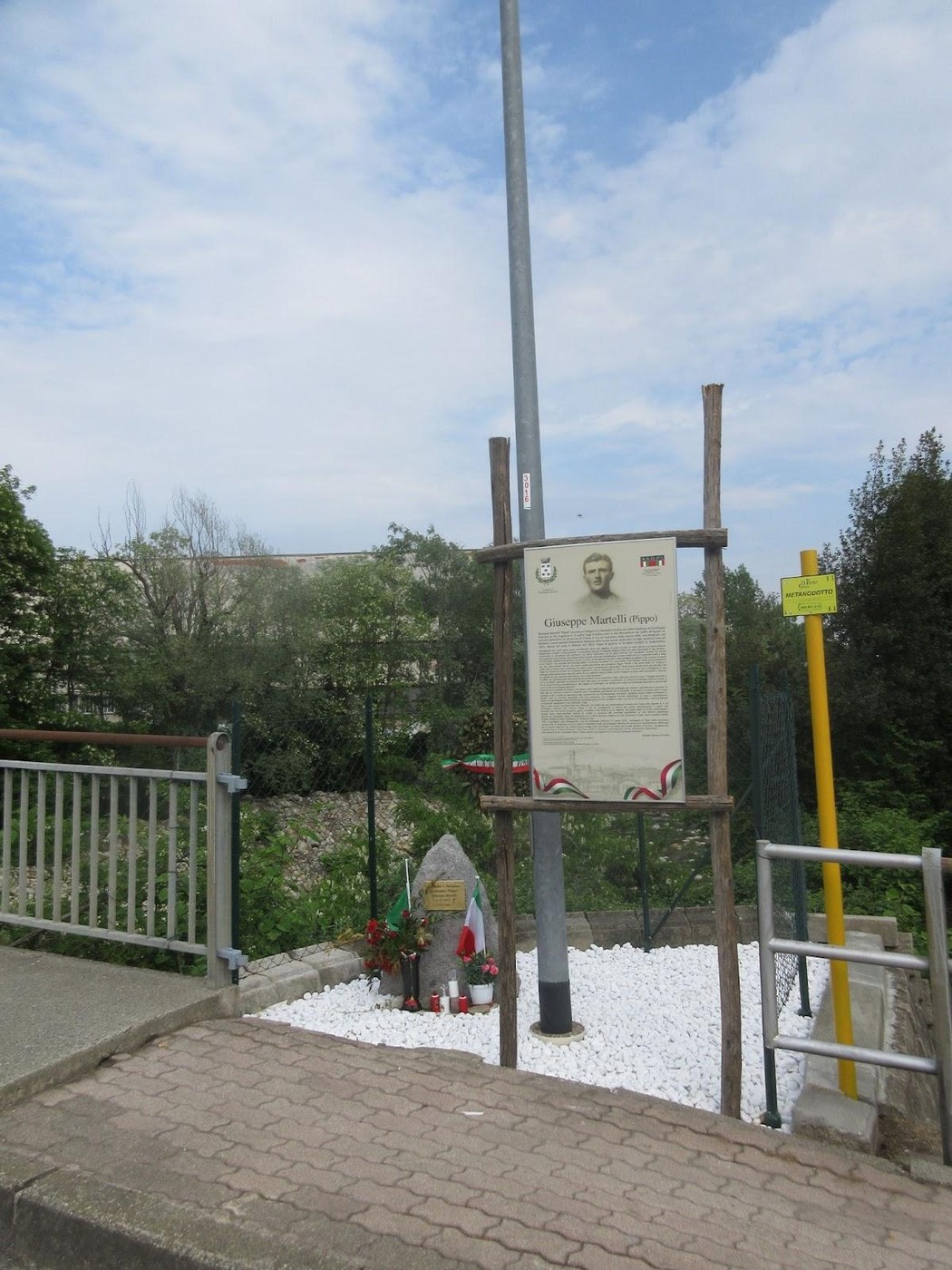 Cippo commemorativo dedicato a Giuseppe Martelli, con fiori e segnaletica in un contesto naturale.
