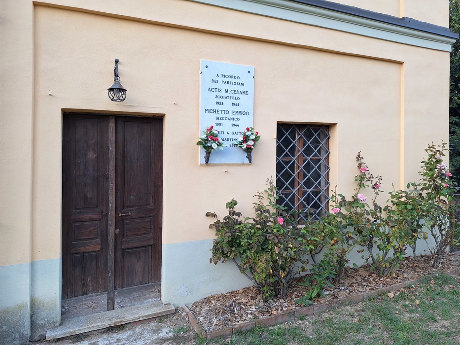 Lapide con iscrizione sulla parete esterna di un edificio, accanto a una porta e a un lampione in ferro.