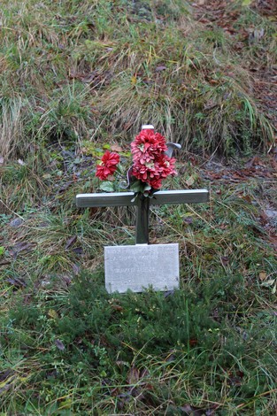 Croce in legno adornata con fiori rossi, posizionata su terreno erboso.