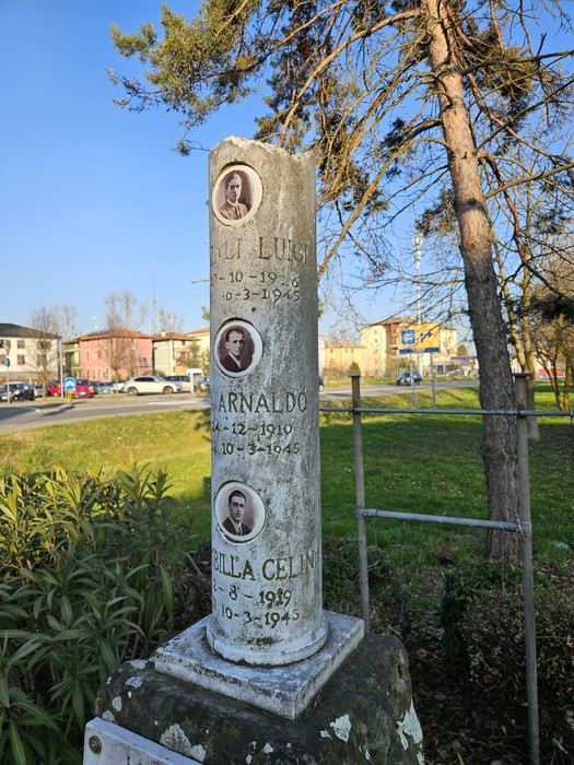 Cippo commemorativo con fotografie di tre persone, situato in un’area verde di una città.