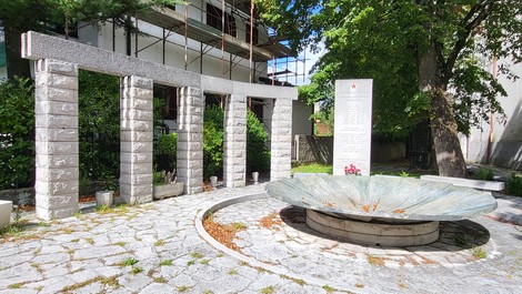 Colonnato in pietra e lapide con iscrizione in un cortile alberato, davanti a una fontana circolare in pietra.