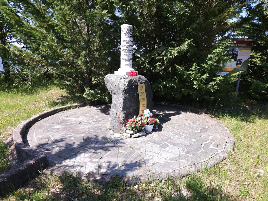 Cippo in pietra con colonna bianca incisa, su base circolare lastricata, circondato da alberi, con fiori deposti ai piedi.